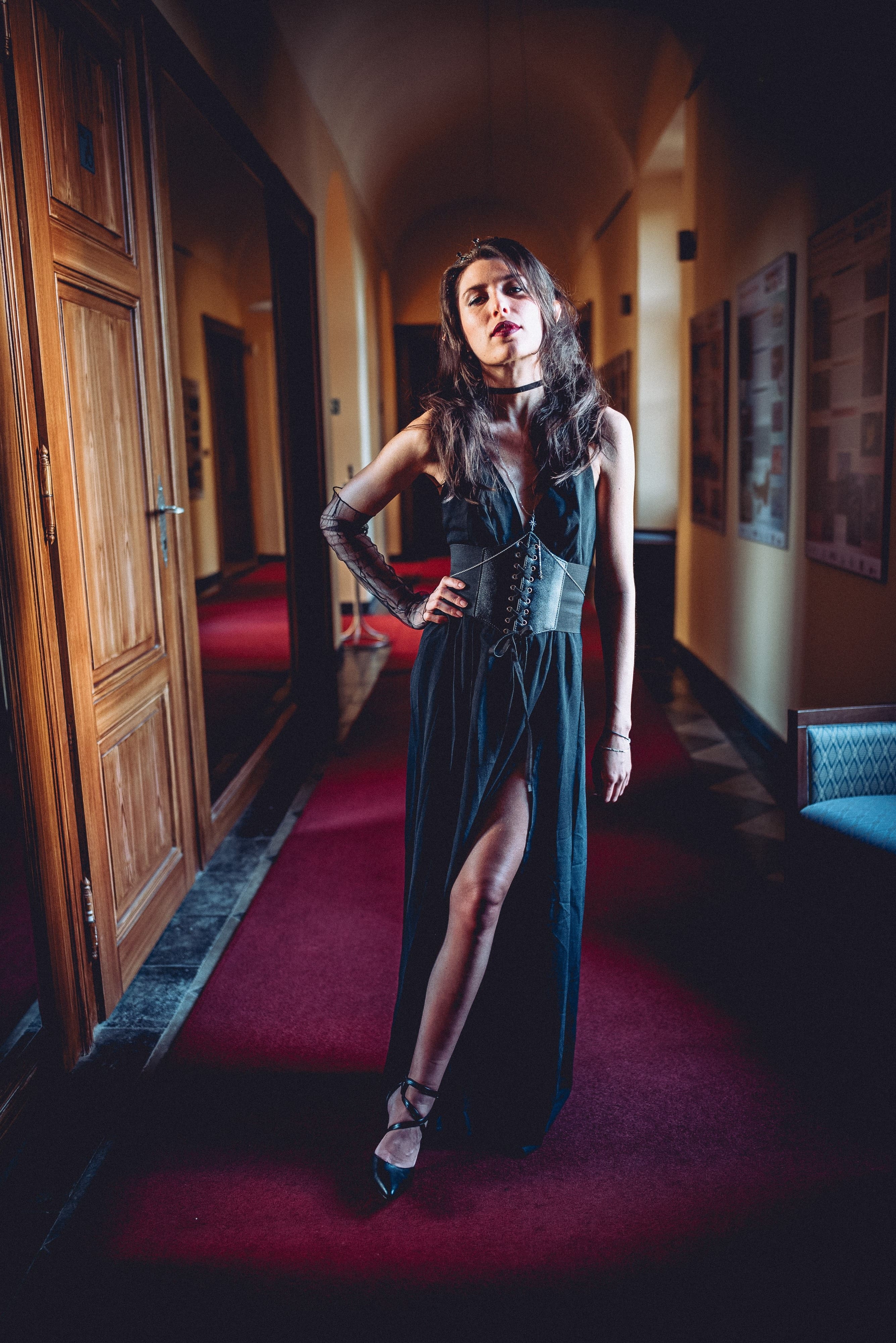 Žena v elegantních tmavých šatech s rozparkem kráčí zámeckou chodbou s červeným kobercem na plese Mystic Star